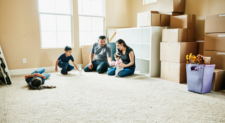The image depicts a family in a room with unpacked moving boxes. The room appears to be well-lit, with light-colored walls and a carpeted floor.